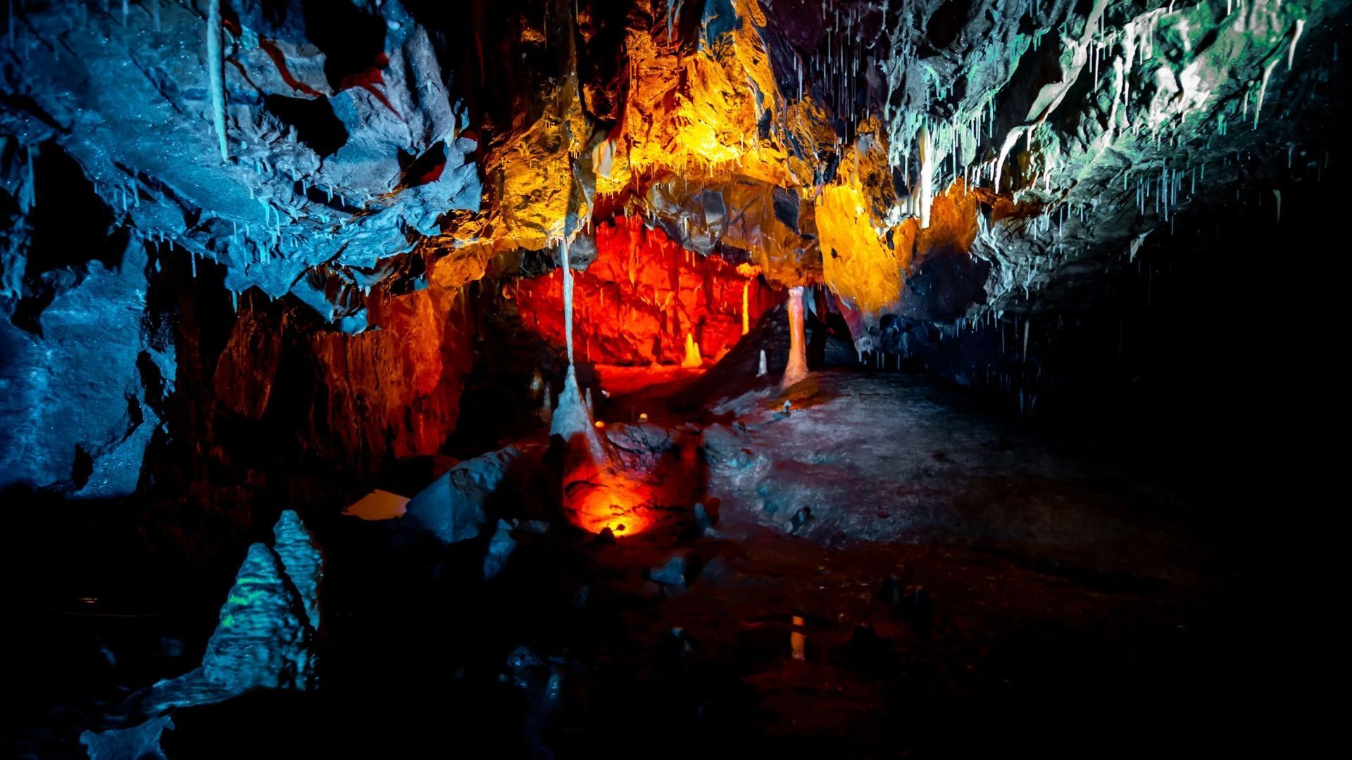 Cave interior with colorful lighting: blue, orange, and red hues illuminate stalactites and rocky formations.