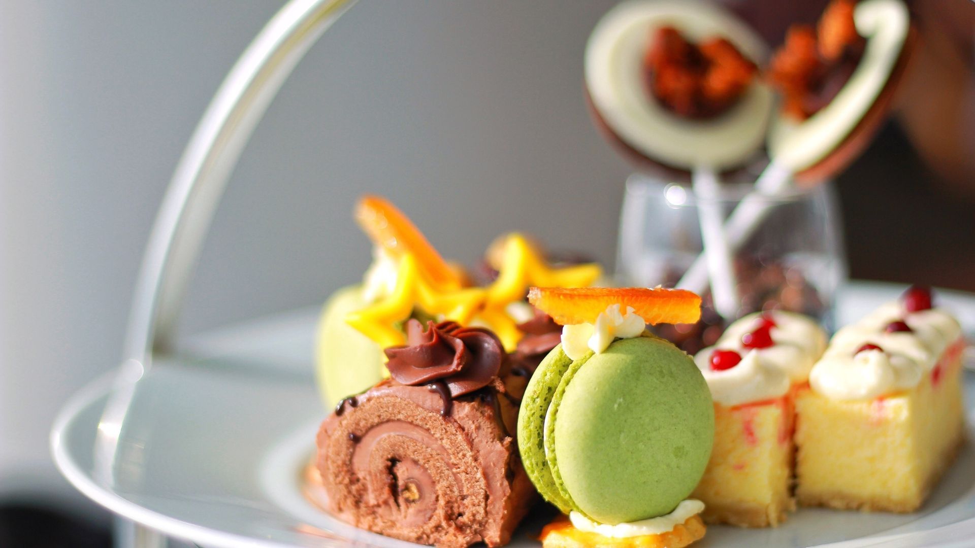 Close-up of a white plate with assorted colorful pastries: a chocolate roll, green macaron, cheesecake, and chocolate lollipop.