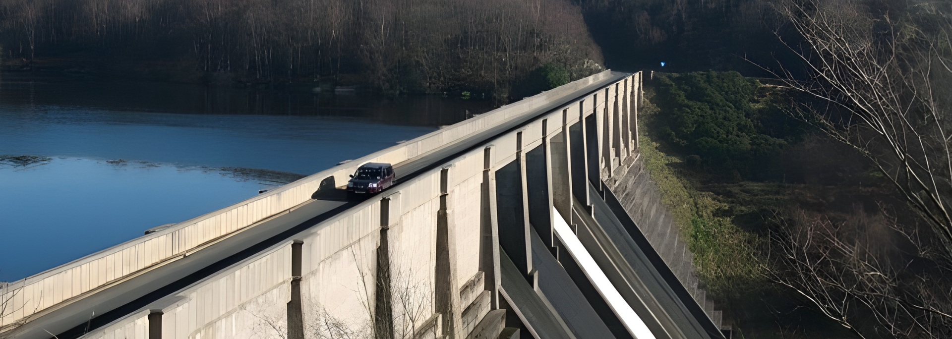 A dark car drives along a dam, with a lake on one side and a wooded area on the other.