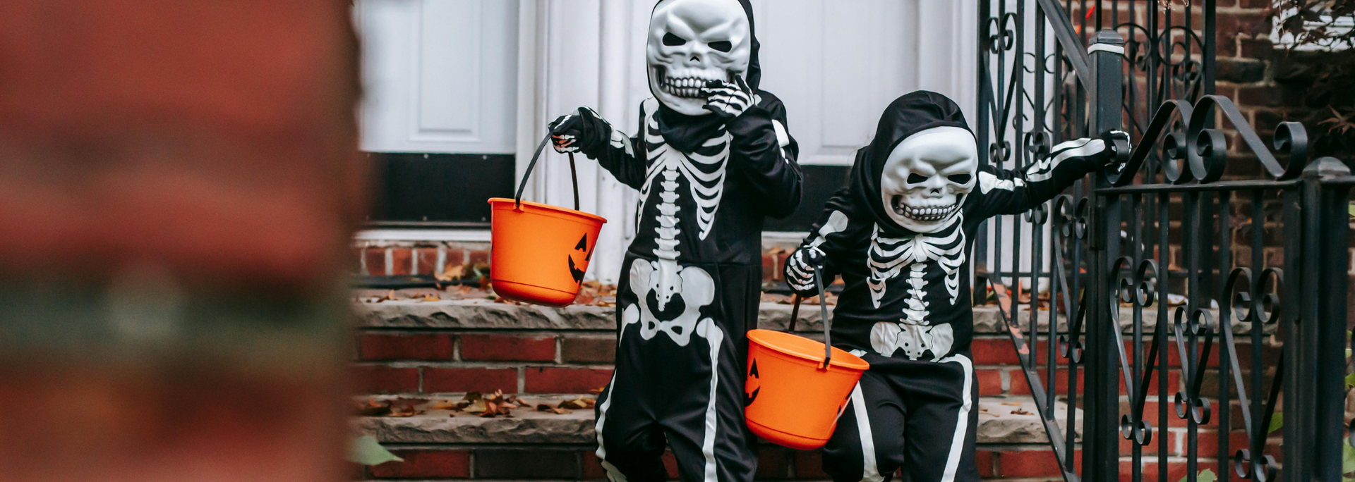 Two children in skeleton costumes holding orange buckets, trick-or-treating on a porch.