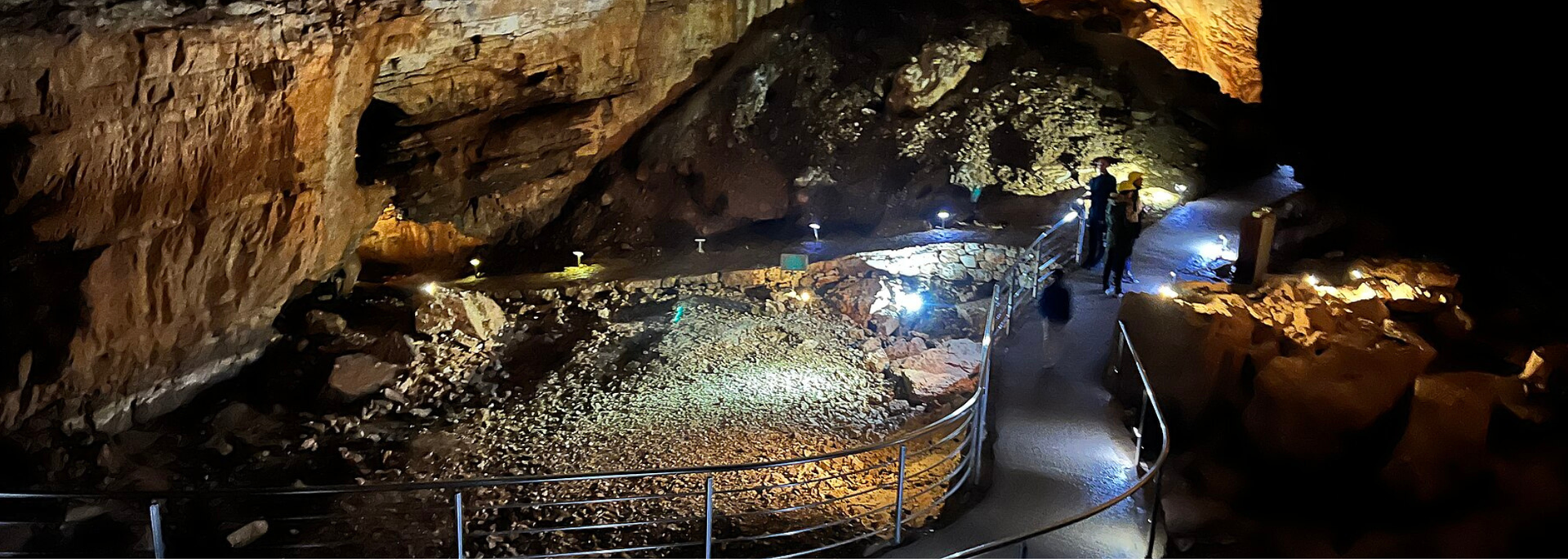 Inside a cave, a pathway illuminated by lights. People walk along the path next to rock formations.