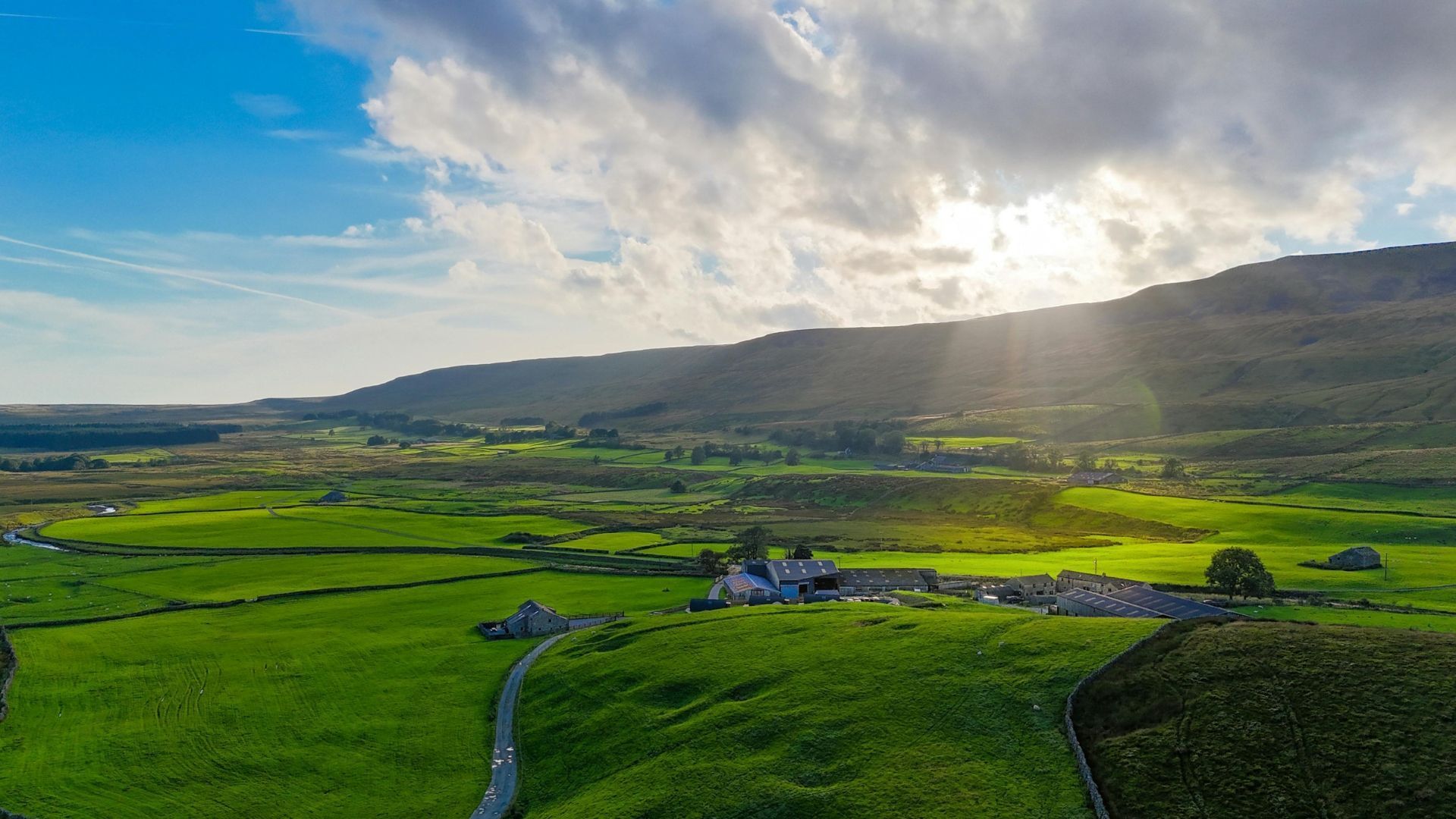 Green valley with a stone building, mountains in background, sun shining through clouds.