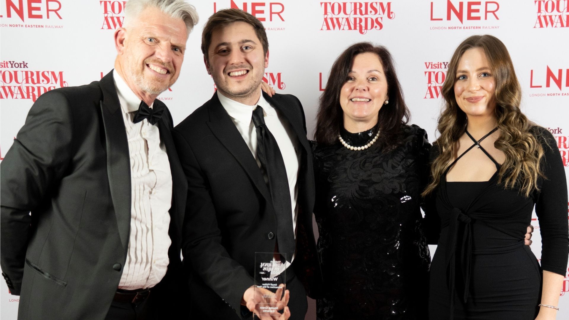 Four people posing with an award at a tourism event. Red backdrop with logos.