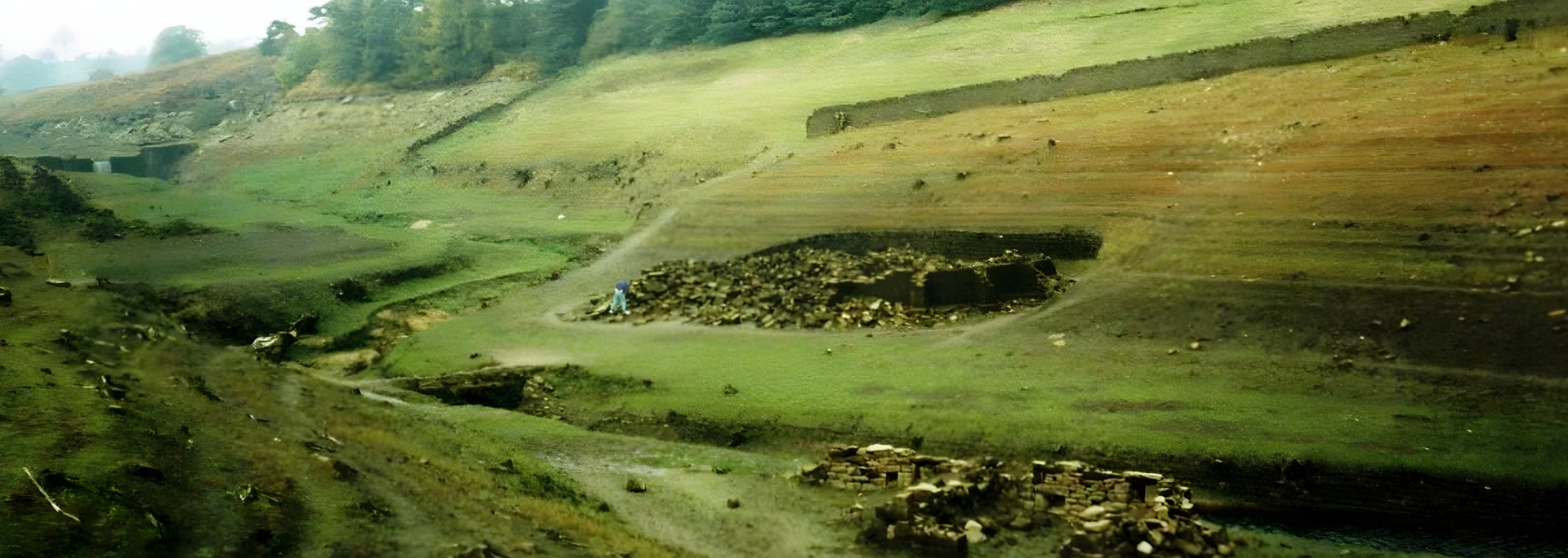 A landscape of a dry riverbed, green grass, and hillside with a stone wall.