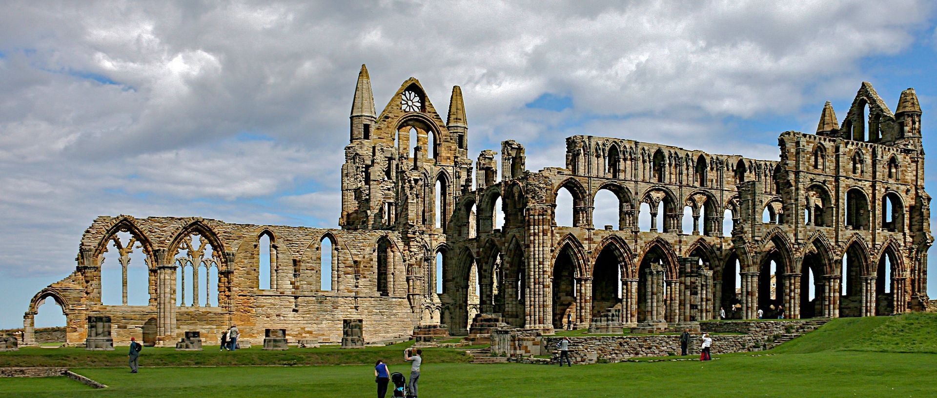Ruined stone abbey on a green hill under a cloudy blue sky. People walk on the grass in the foreground.