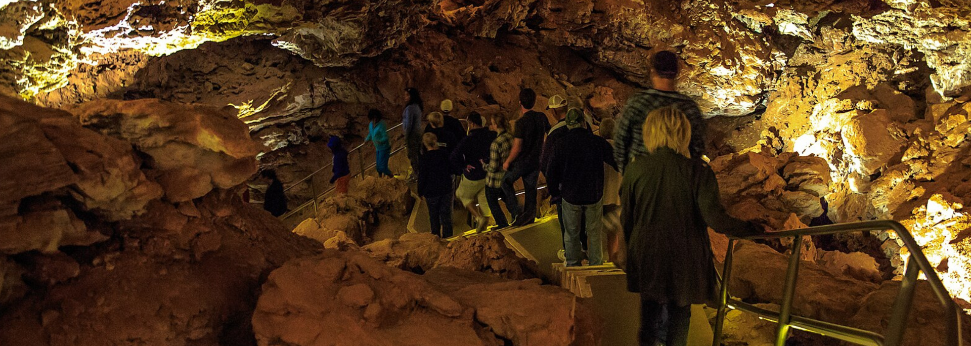 People walking through a cave, illuminated by lights. The cave walls are rocky and brown.