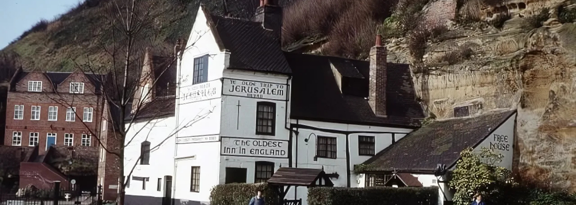 White timber-framed building nestled into a hillside, with a dark roof and chimney, and another building in the background.