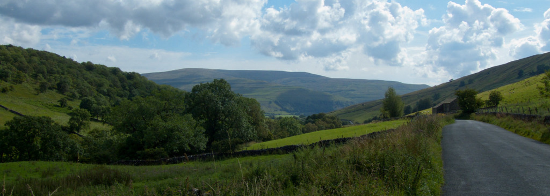 Rolling green hills under a cloudy sky. A road winds through a valley.
