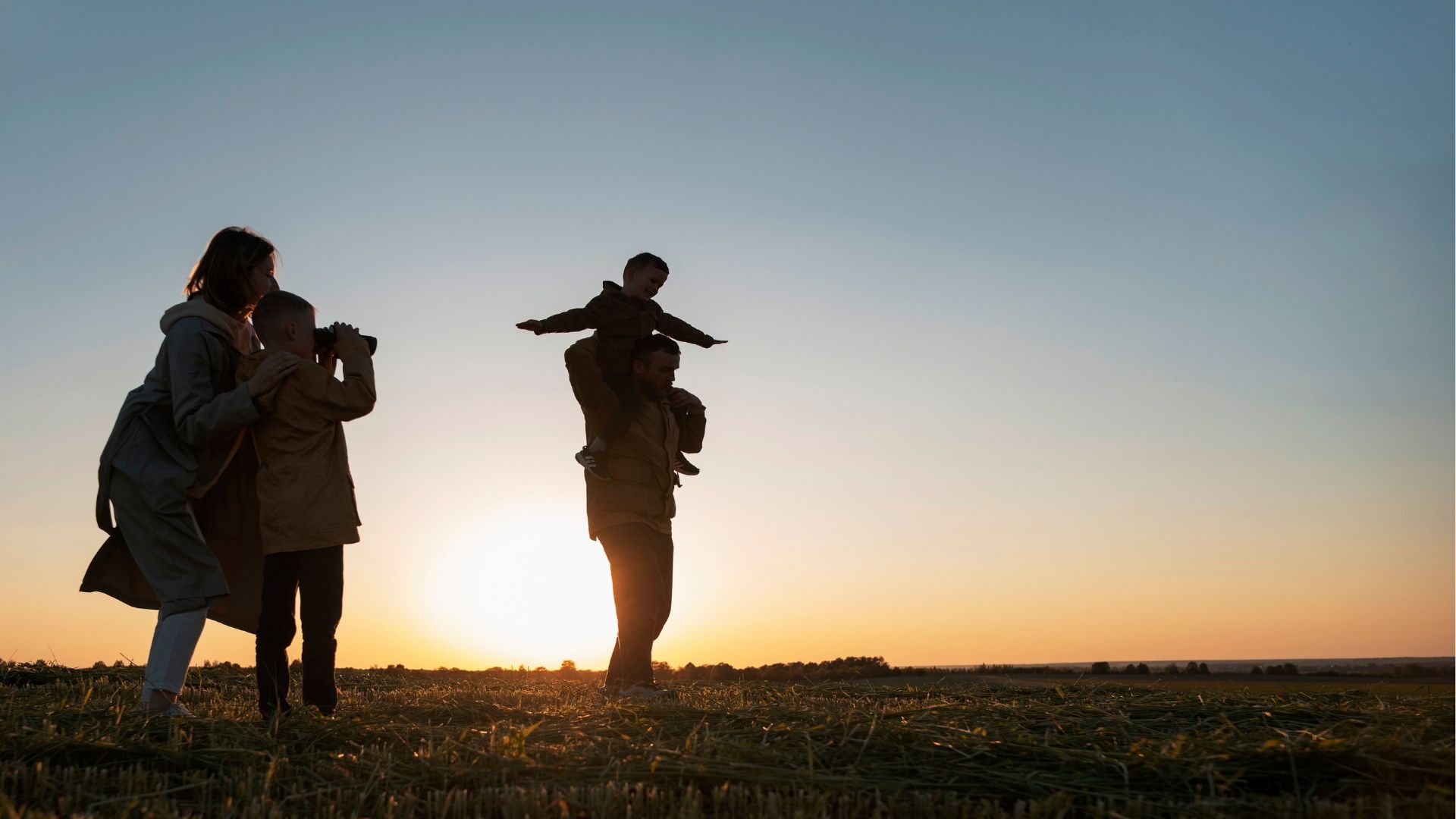 Family silhouetted in a field at sunset: father carrying child on shoulders, others looking on.