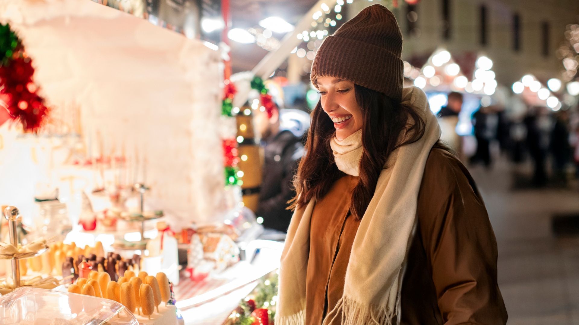 Woman in winter clothes smiles at a holiday market stall, lit by festive lights.