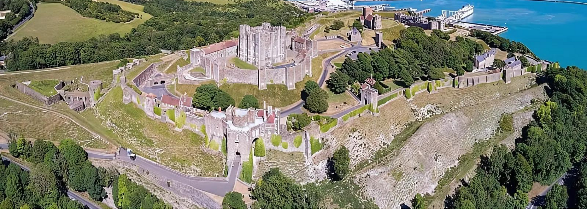 Aerial view of a castle on a hill with a blue sea in the background. Green trees surround the castle.