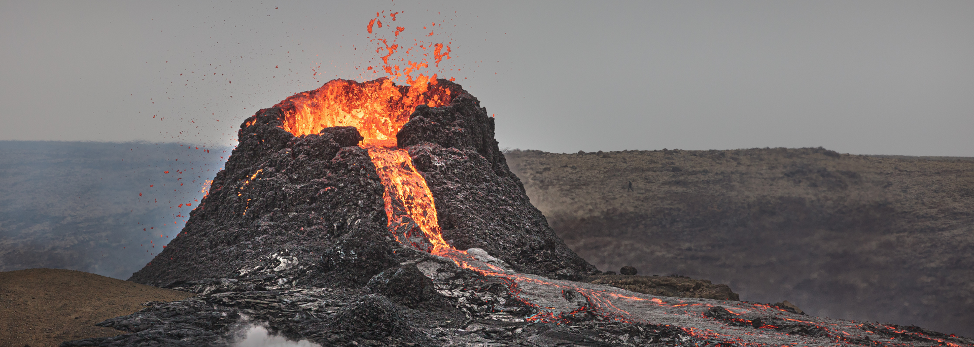 Volcano erupting, spewing lava. Bright orange magma against a gray sky, flowing over dark volcanic rock.