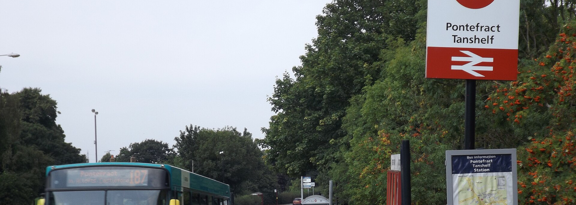 A train station sign, with the British Rail symbol, next to a bus and greenery under an overcast sky.