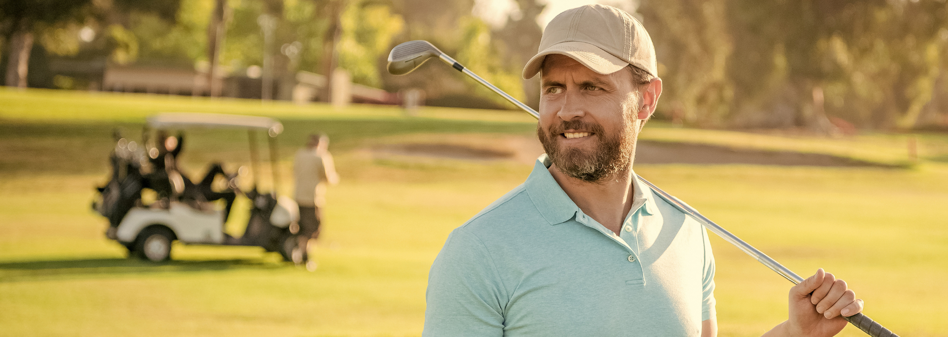 Man on golf course with club over his shoulder, smiling. Golf cart in background, sunny day.