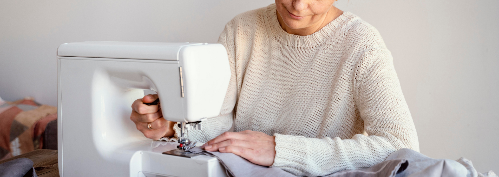 Person in cream sweater using a white sewing machine, working on fabric. Bright setting.