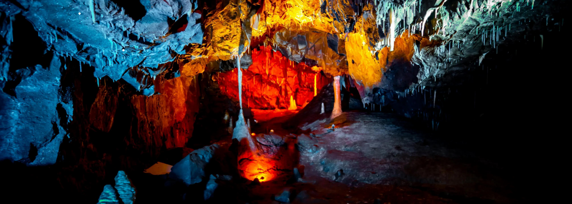 Inside a cave, red and orange light illuminate formations on walls and a reflecting pool.