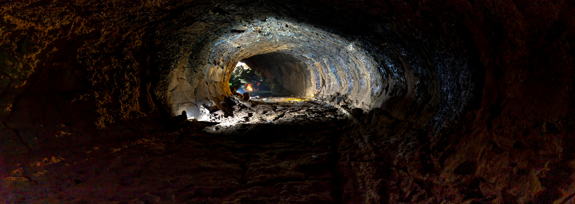 Inside a dark cave, a tunnel with illuminated opening, possibly volcanic.