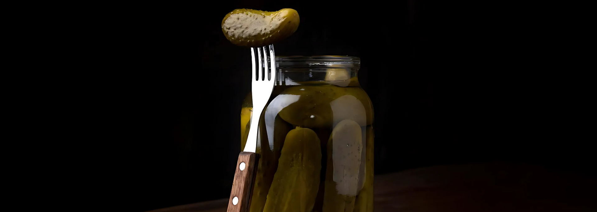 A jar of pickles with a slice impaled on a fork. Dark background.