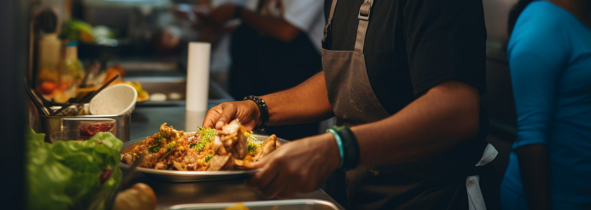 Person holding a plate of food in a busy kitchen. Tacos are on the plate.