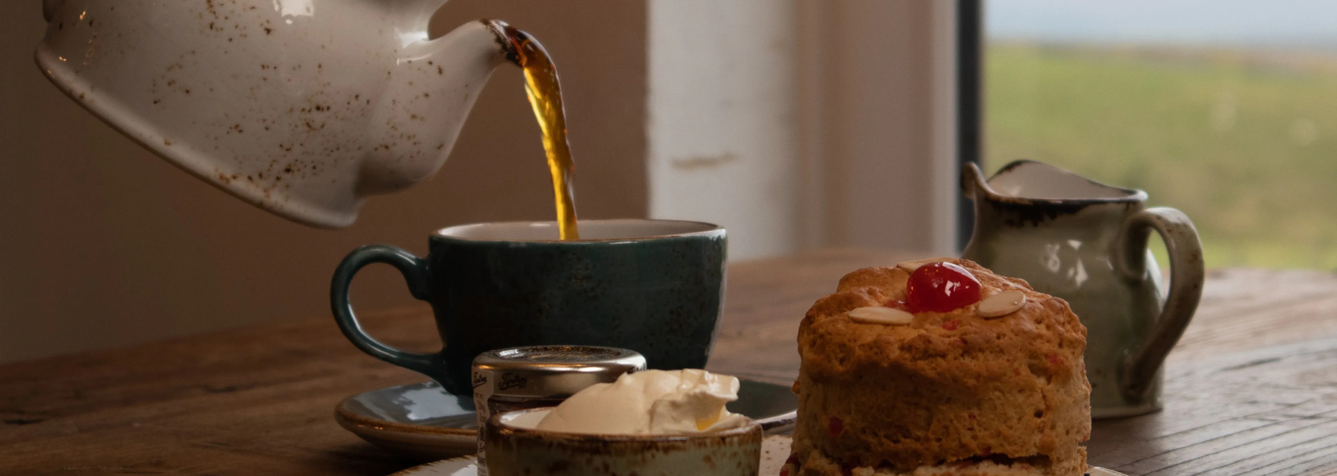 Tea being poured into a teacup with a scone and cream on a wooden table near a window.
