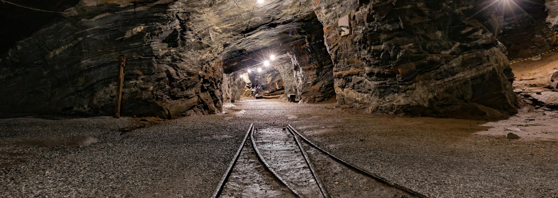 A mine tunnel with tracks. Dim lighting reveals stone walls and ceiling; lights illuminate the path ahead.