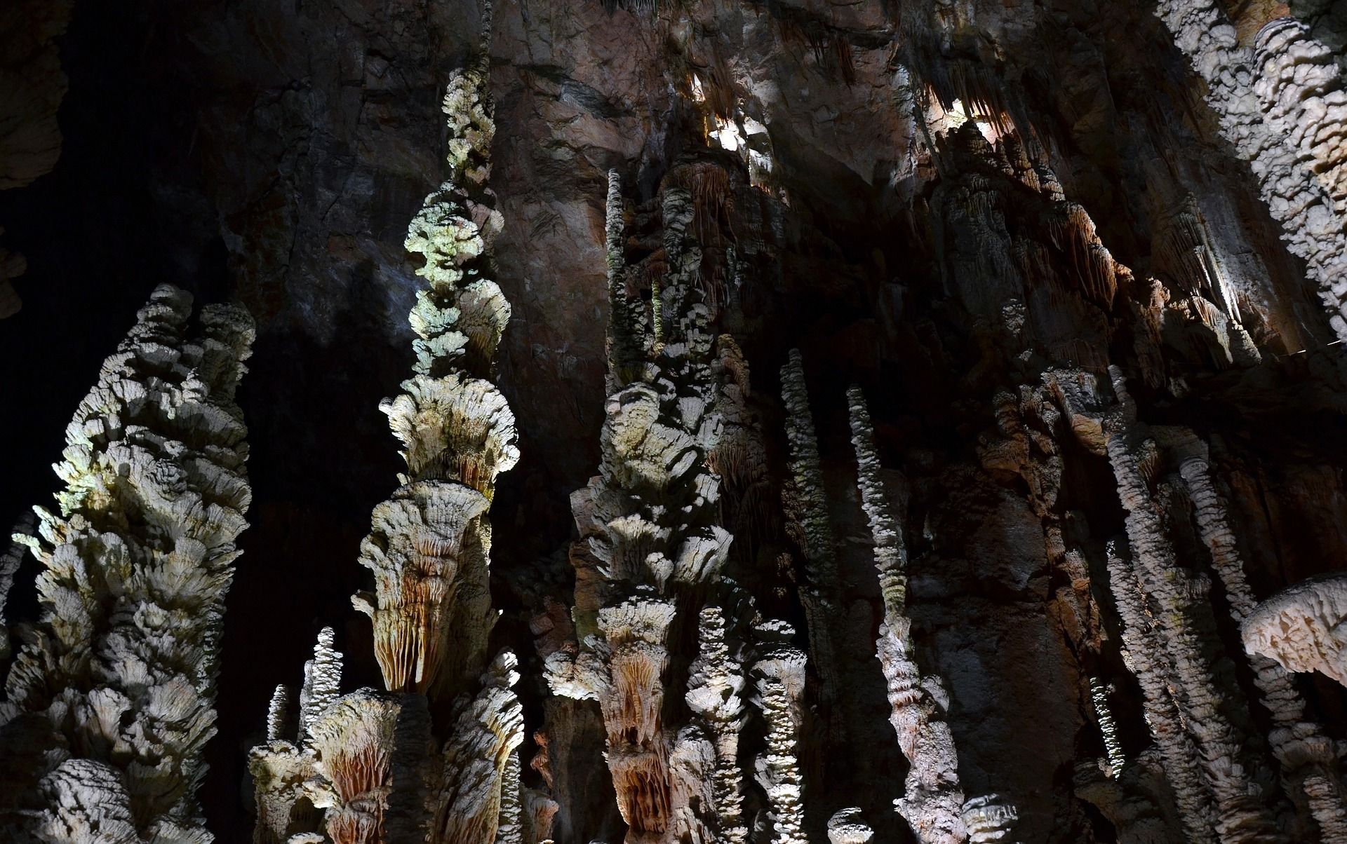 Cave interior, illuminated stalactites and stalagmites in varying sizes and textures, dark backdrop.