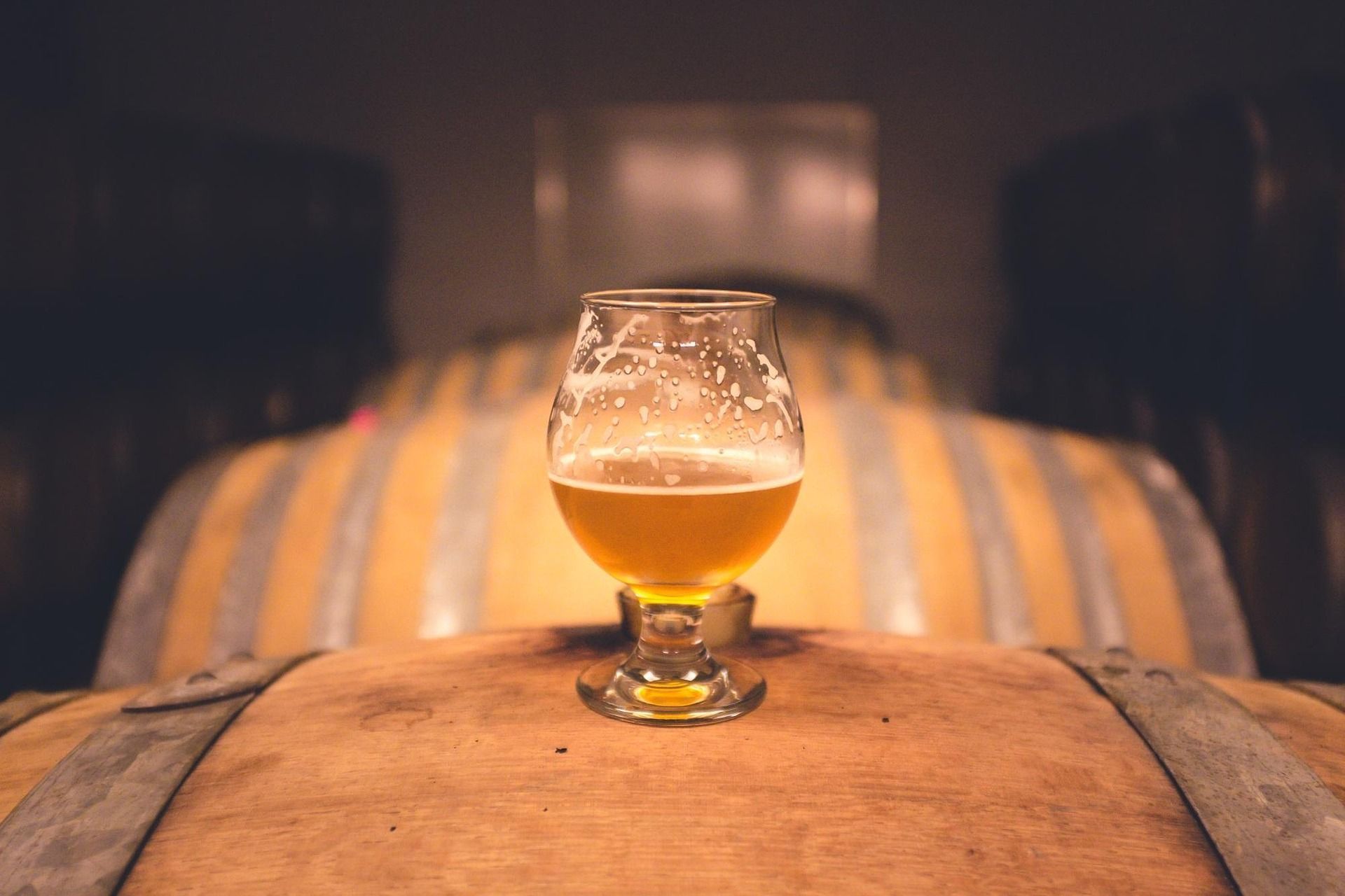 Glass of beer on a wooden barrel, in a brewery, with blurred barrels in background.