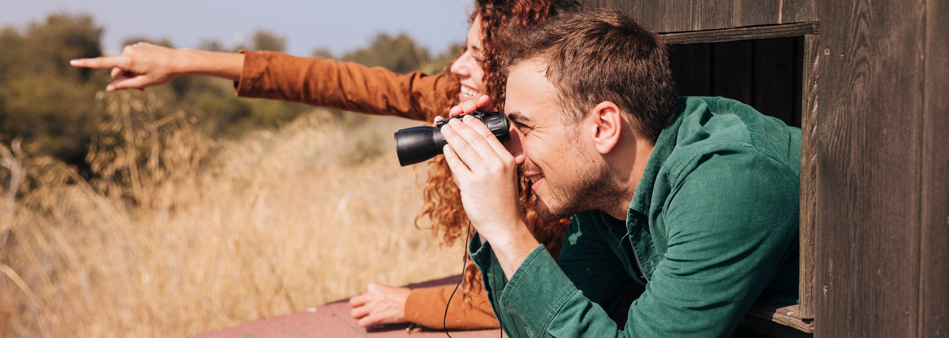 A person looks through binoculars, another points. They are in a wooden structure overlooking tall grass.