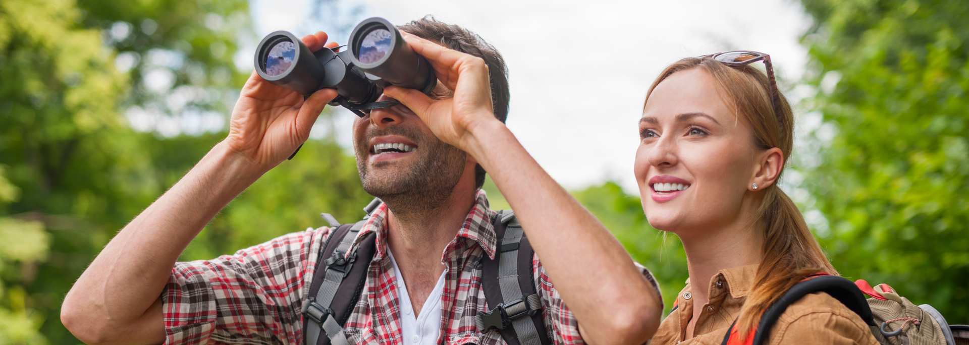 Man using binoculars, woman looks on. Both smile, enjoying nature, surrounded by trees.