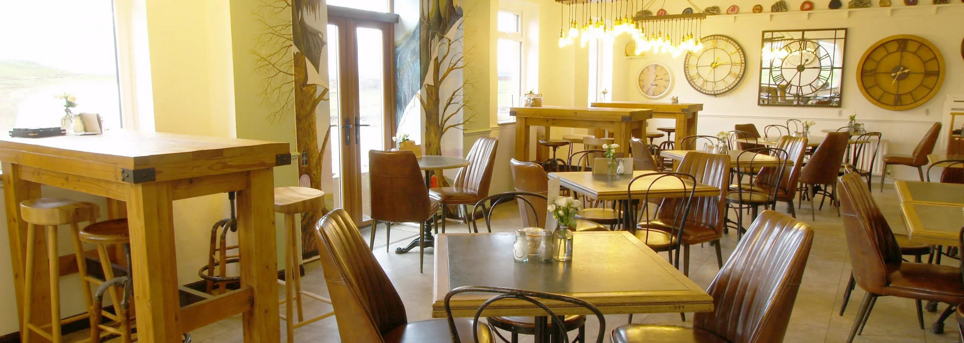 Interior view of a cafe with wooden tables and chairs, decorative clocks on the wall, and natural light.