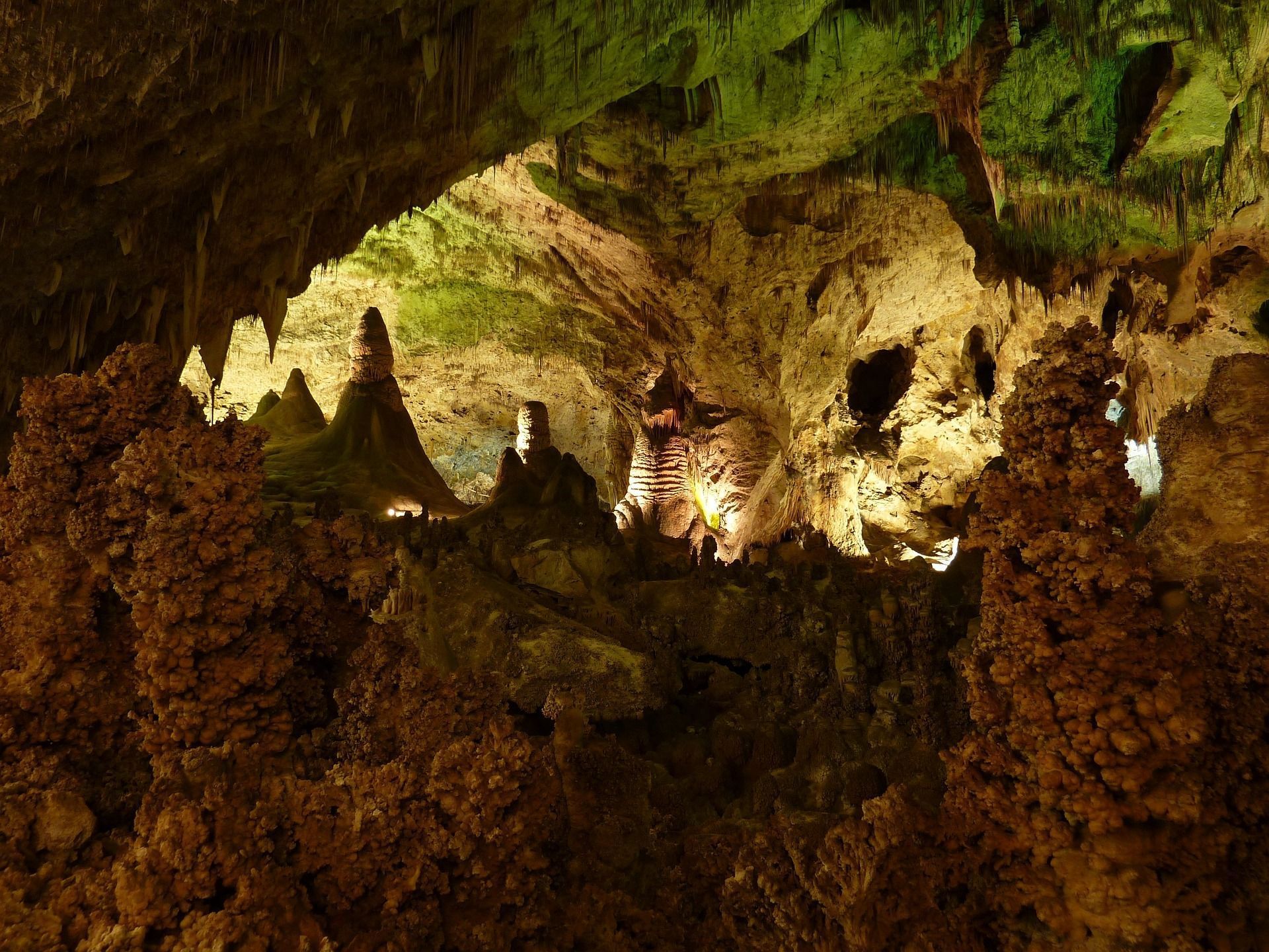 Cave interior with illuminated stalagmites and stalactites; earthy tones, green highlights.