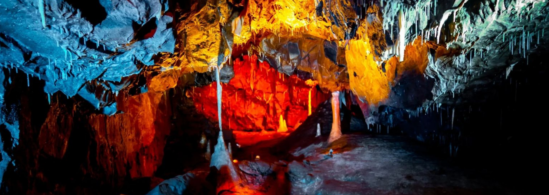 Cave interior illuminated with blue, orange, and red lights, showcasing stalactites and geological formations.