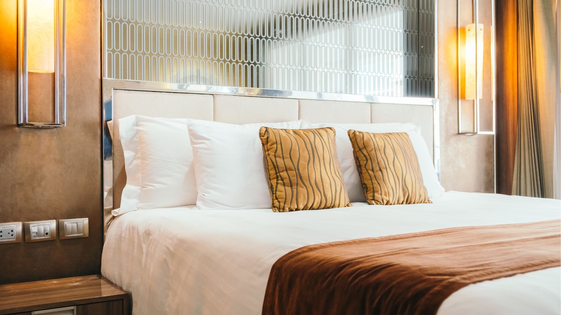 Bedroom with a bed, white pillows, brown patterned throw pillows and blanket, glass headboard, bedside table, and sconces.