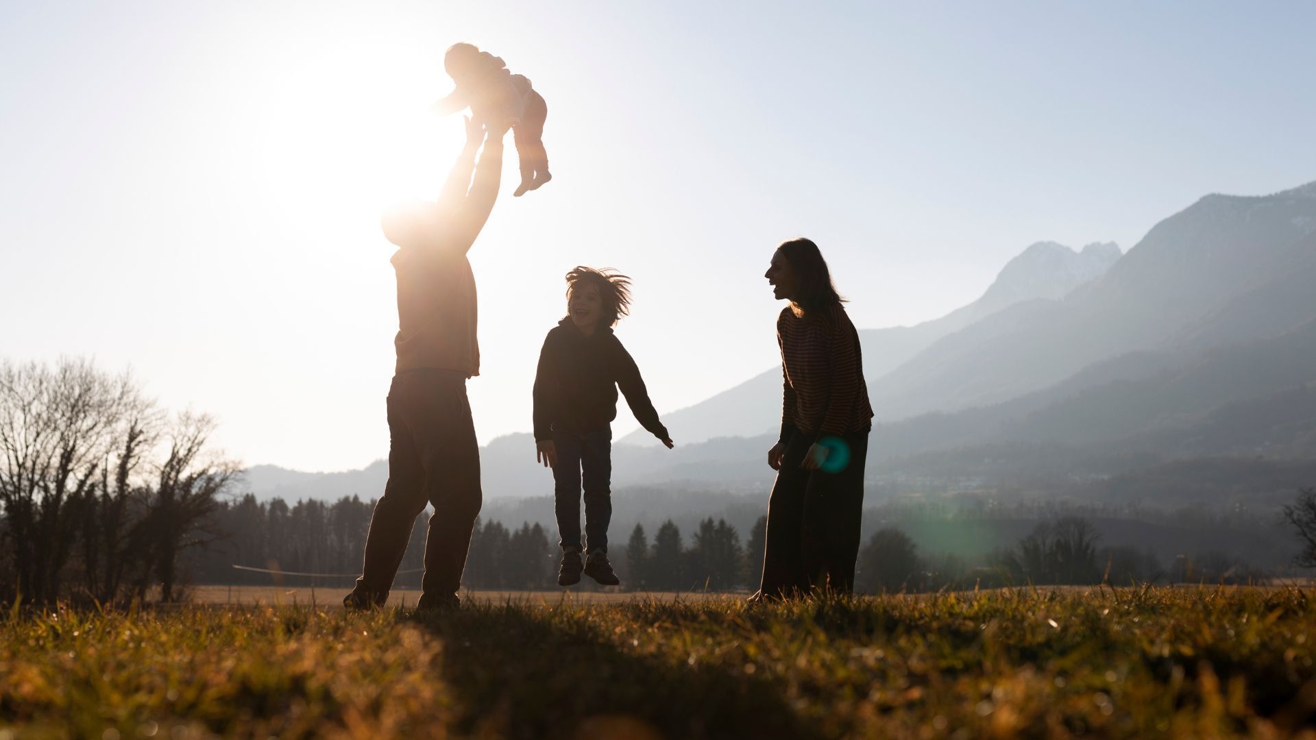 Family playing outdoors with mountain backdrop; parent holding child up, another child jumping.
