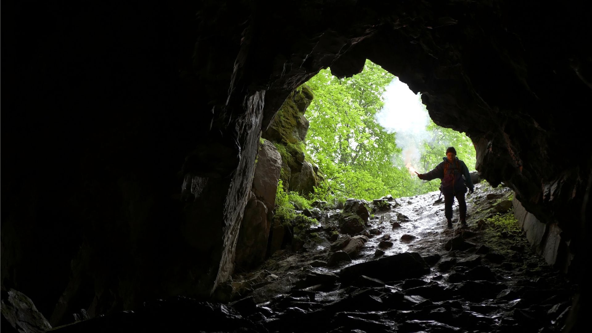 Person exiting cave into a bright, green landscape; dark interior, wet ground, silhouette.
