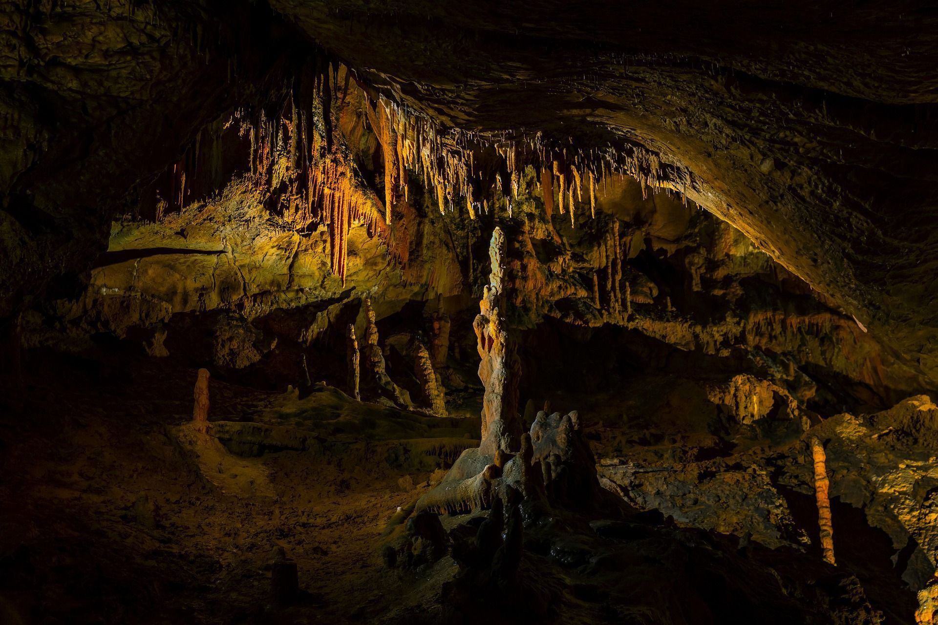 Cave interior with brown stalactites and stalagmites, illuminated with yellow light.