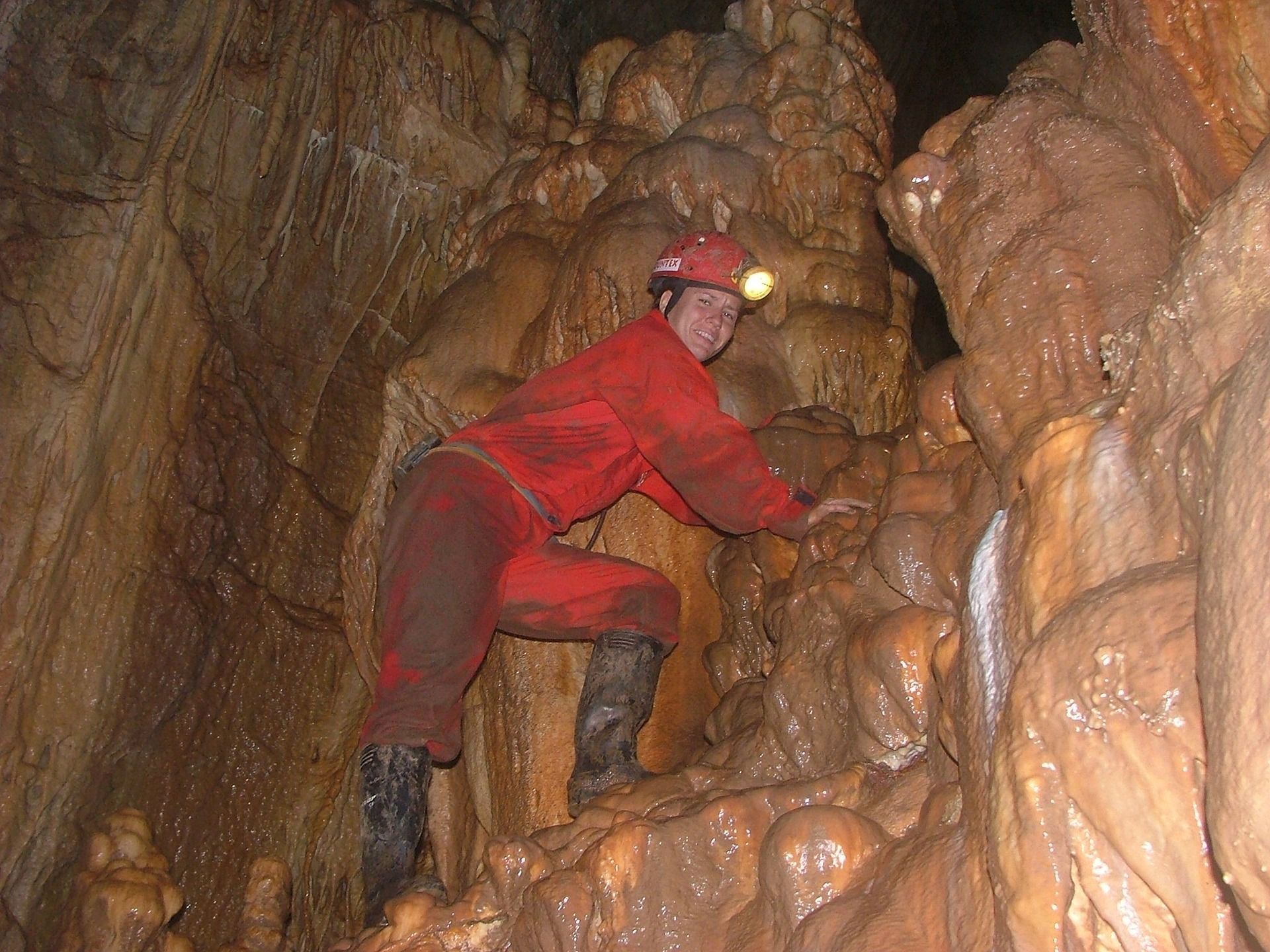 Person in red jumpsuit climbs in a brown cave, wearing a helmet with a headlamp.
