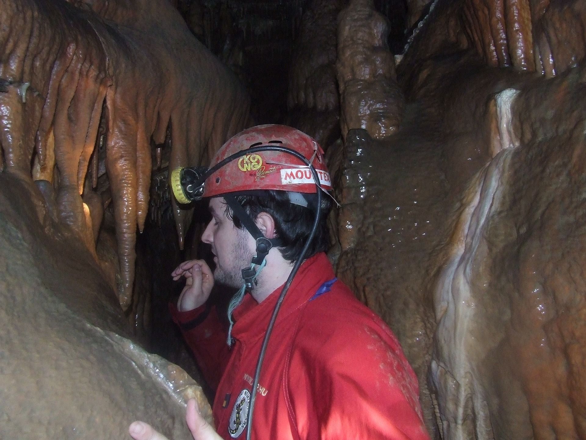 Person with a headlamp and red suit inside a cave, surrounded by rock formations.