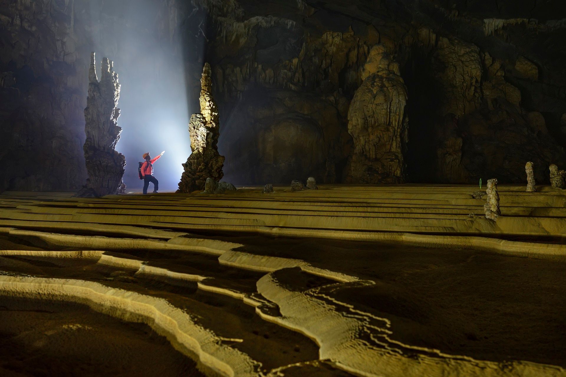Person standing in a large cave, holding a light toward an opening with bright light. Rocky formations and pools.