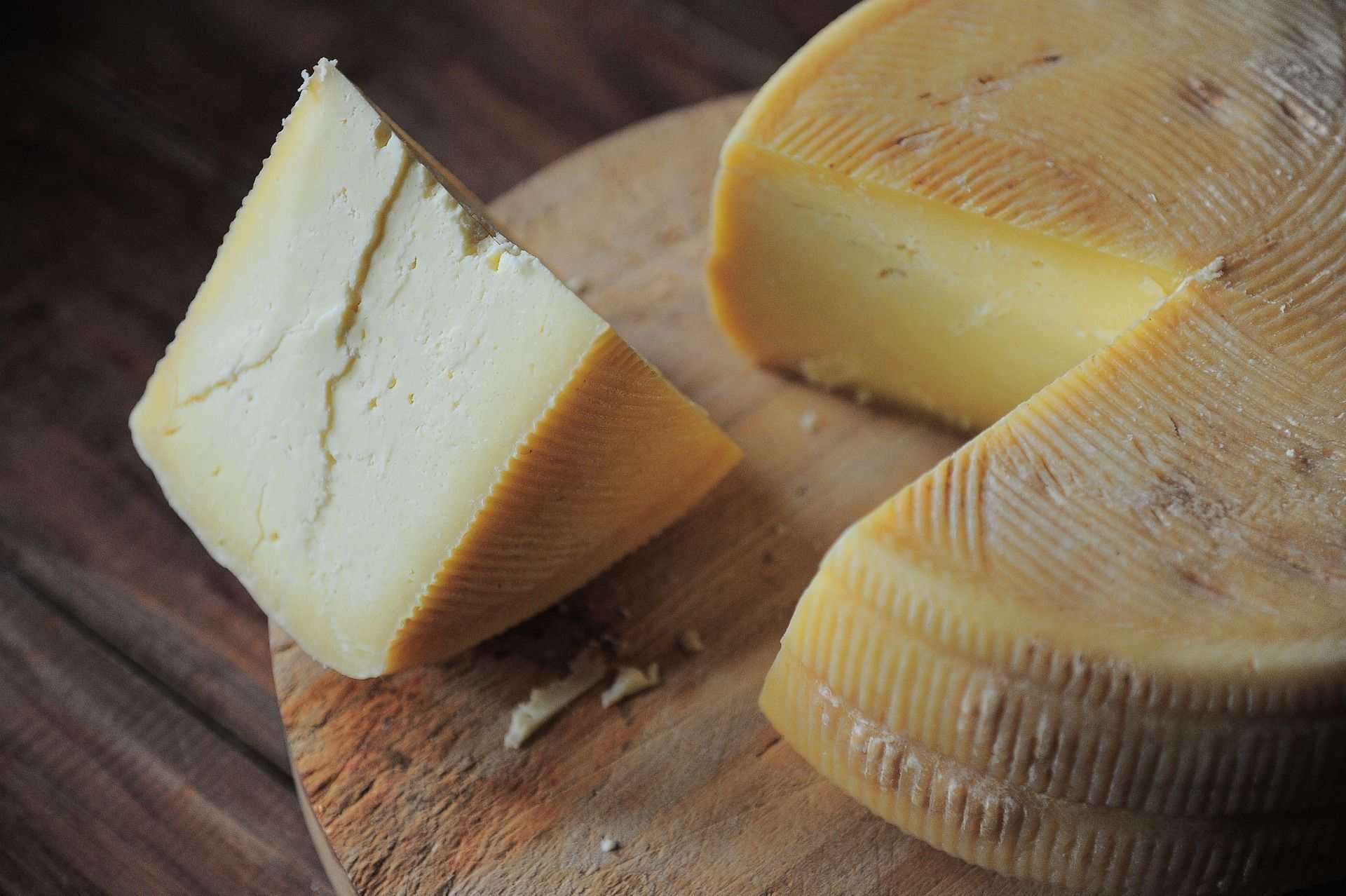 Cheese wedge on a wooden cutting board, with a partially cut wheel in the background.