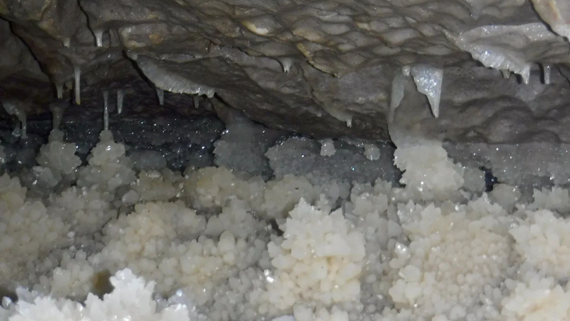 Inside a cave, formations of white crystals hang from the ceiling and grow upwards from the ground.