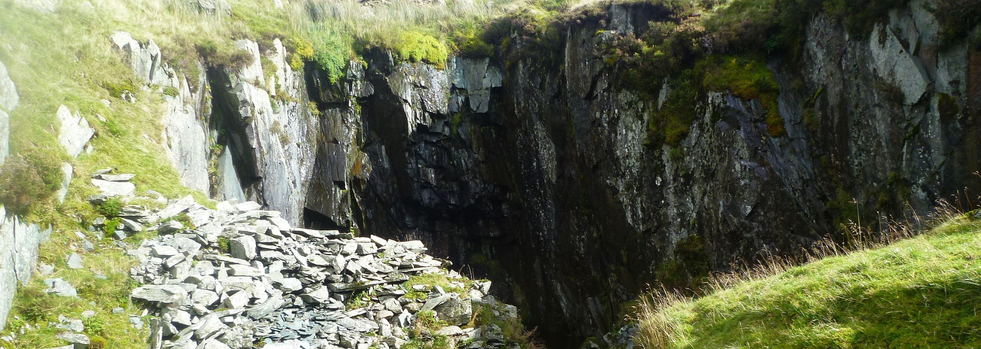 A deep, rocky ravine with patches of green grass and vegetation, weathered gray stone.
