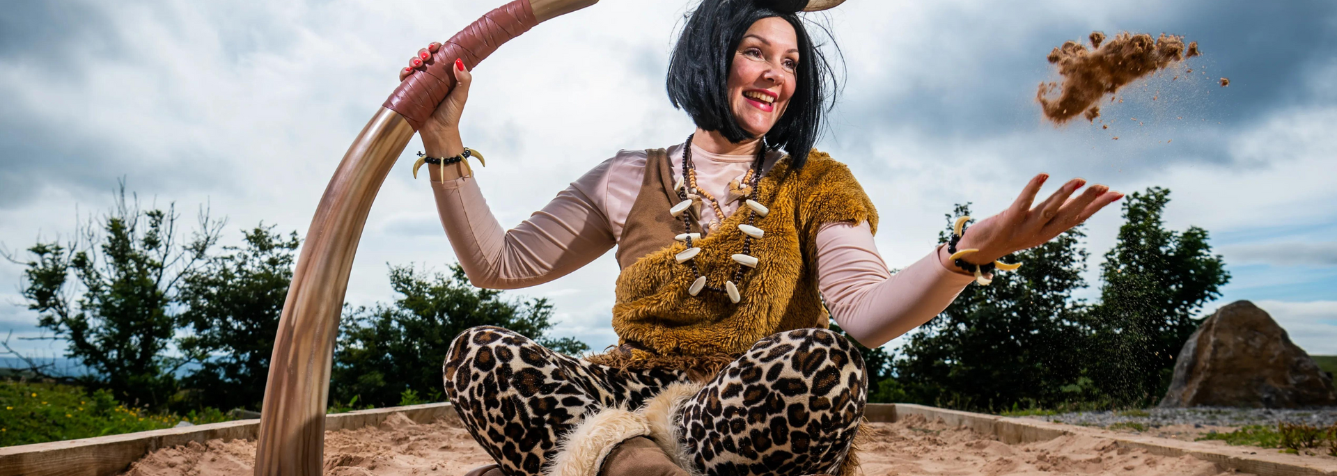 Woman in cavewoman attire smiles, throwing dirt in the air while holding a large club.
