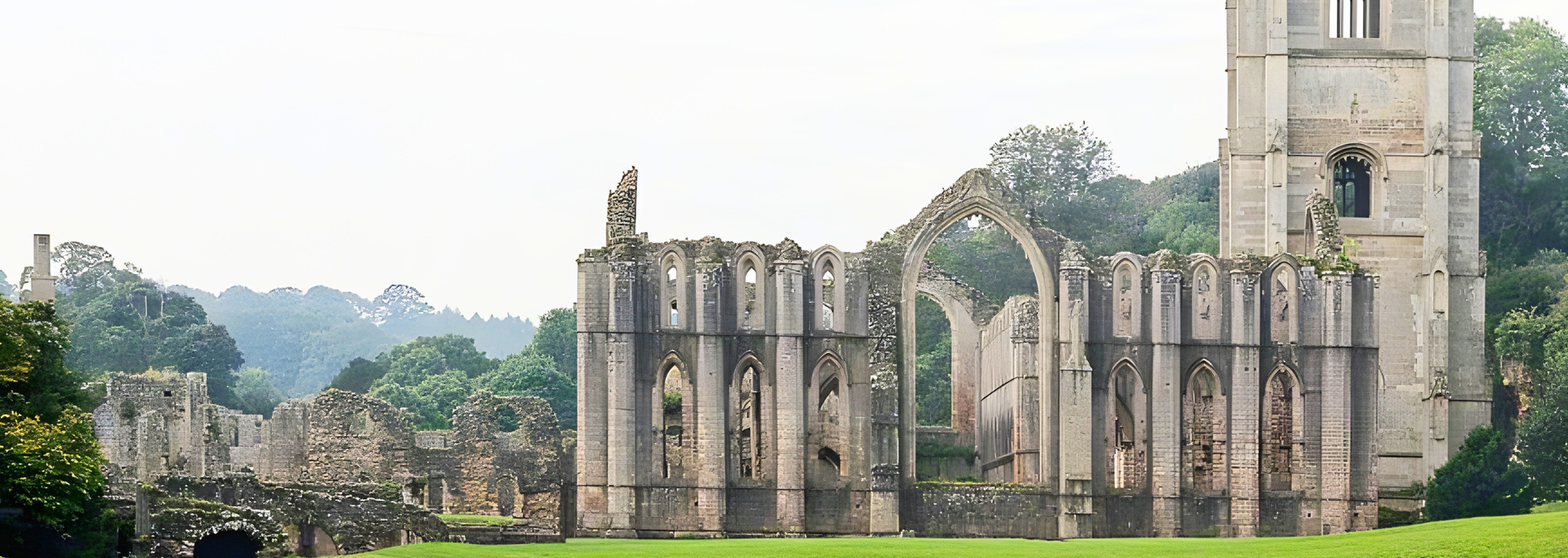 Ruined stone abbey on a green lawn, with a tall tower. Lush green trees and a cloudy sky in the background.