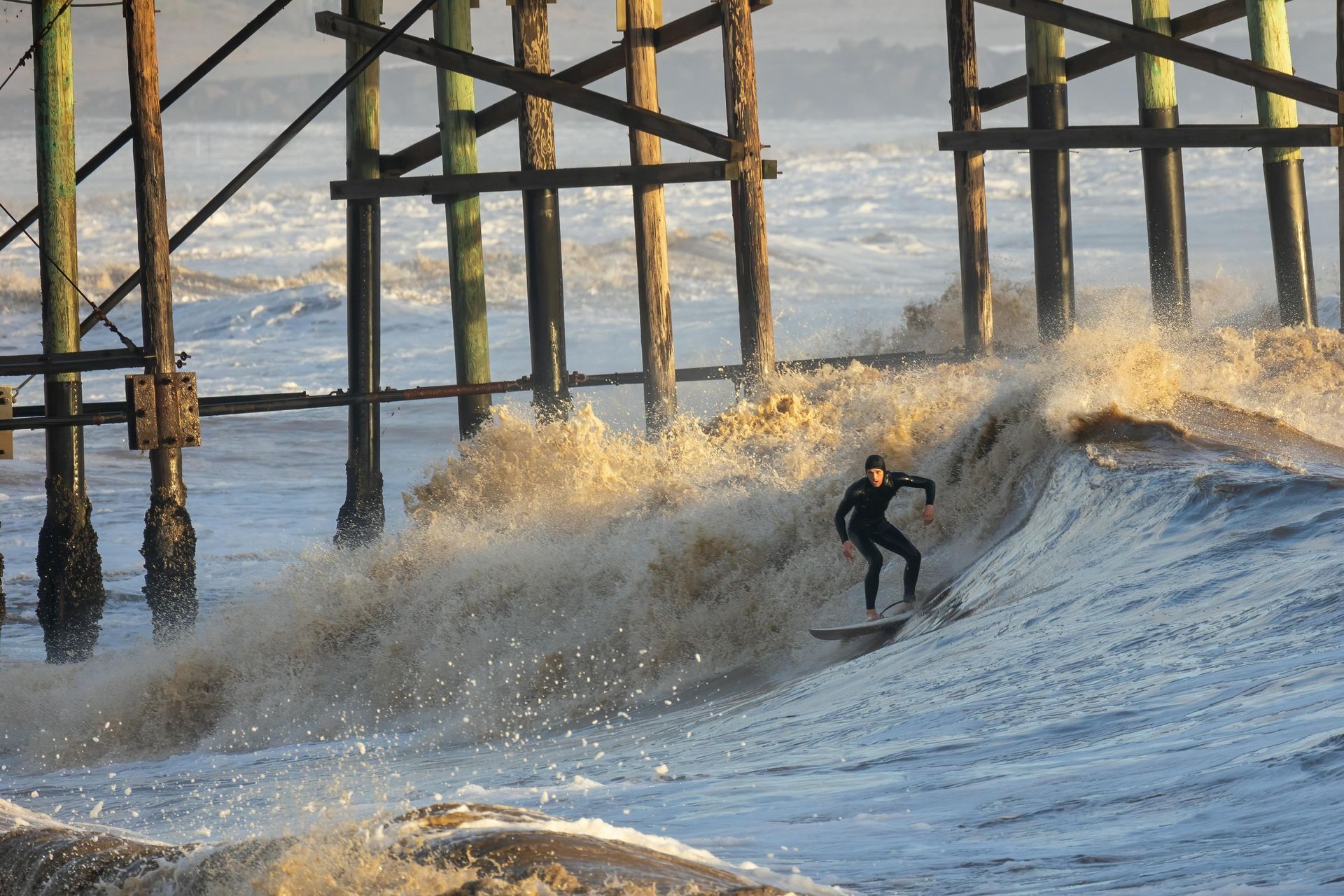 Surfer rides a wave under a wooden pier; water is foamy and gold-toned from sunlight.