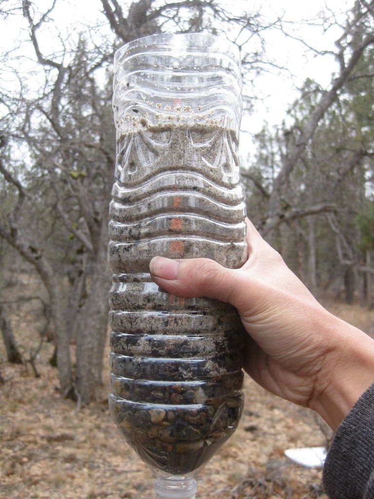 A hand holding a clear plastic bottle filled with layers of soil and rocks outdoors.