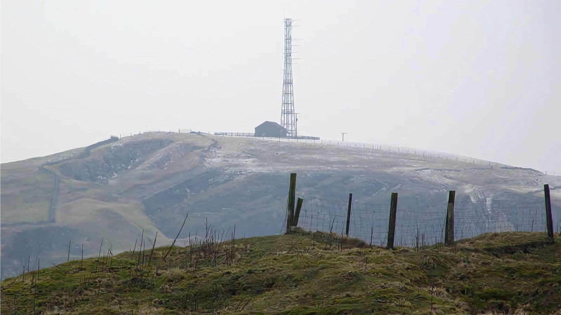 Hilly landscape with a tall radio tower on a snow-dusted peak, fence in foreground.