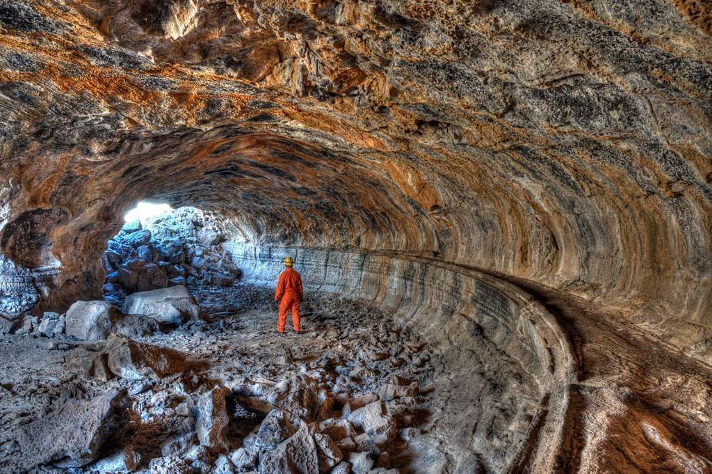 Person in orange suit stands in a lava cave, sunlight at the opening.