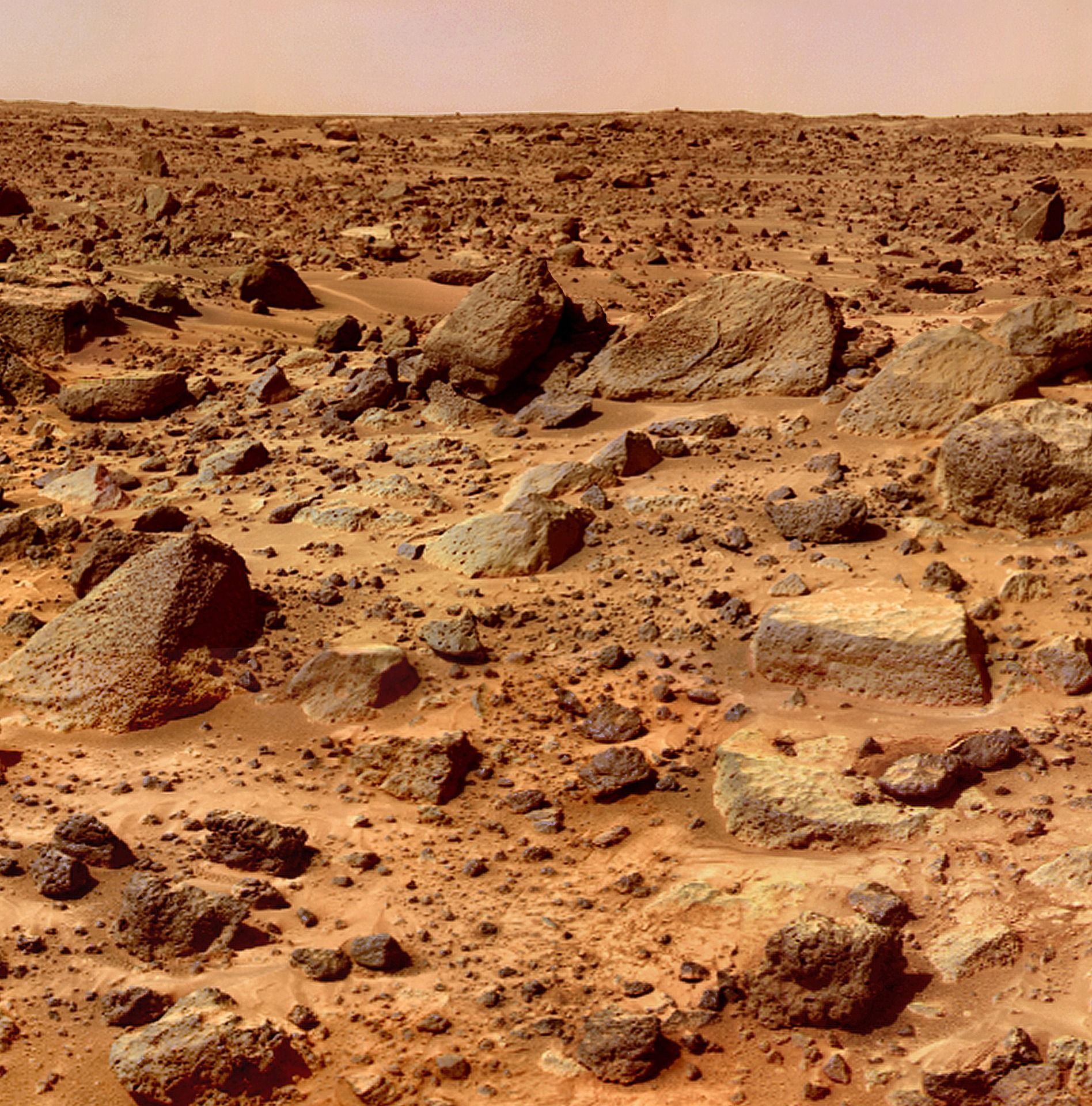 Mars landscape with rust-colored rocks and reddish soil. Horizon and hazy, orange sky visible.