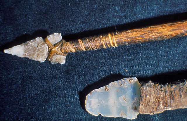 Close-up of a wooden arrow with a stone arrowhead, secured with twine, resting on a dark surface.